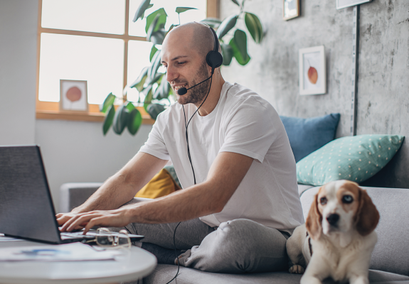 man sitting on couch, working on laptop, with headset on and dog next to him