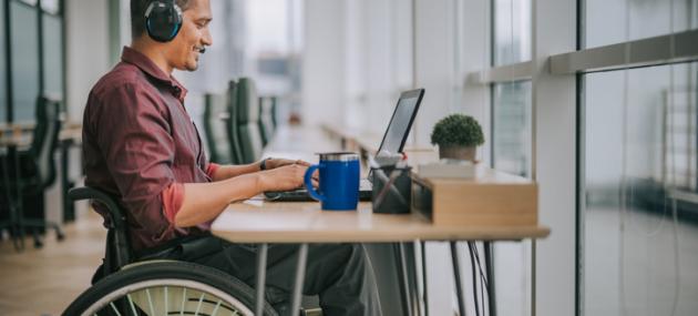 man in wheelchair sitting at desk with headphones on, working on laptop