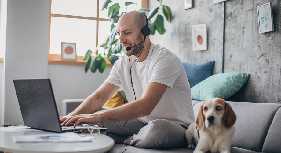 man sitting on couch, working on laptop, with headset on and dog next to him